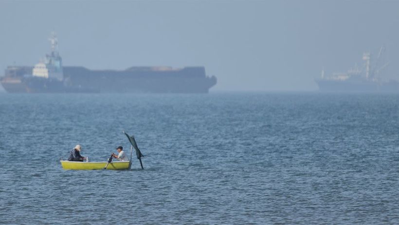 Two men sit in a small boat on the water as cargo ships are anchored in the background in the Strait of Hormuz off Bandar Abbas, Iran, Monday, May 4, 2026. (Amirhosein Khorgooi/ISNA via AP) XEM102